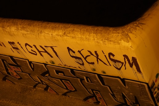 The image depicts graffiti art on a concrete wall. The words 'FIGHT SEXISM' are spray-painted in bold, dark letters, while below them there is another section of graffiti with abstract shapes and letters. The wall and the surrounding area appear weathered and the lighting gives a yellowish hue to the scene.