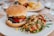 A sesame seed bun burger with lettuce, tomato, and a patty has a knife inserted vertically into it. Next to the burger is a colorful quinoa salad with vegetables such as chickpeas, red peppers, corn, and greens. The meal is served on a white plate with a leaf design, with a blurred background showing another plate of food.