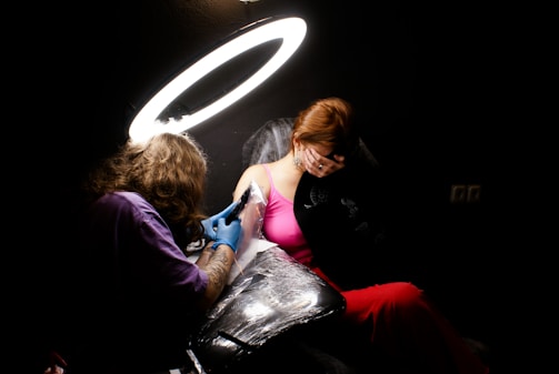 A tattoo artist with long hair is working on a client's shoulder under a bright ring light. The client, wearing a pink top and red skirt, appears to be shielding their face with one hand. The room is dimly lit, creating a strong contrast between the lit area and the dark surroundings.