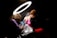 A tattoo artist with long hair is working on a client's shoulder under a bright ring light. The client, wearing a pink top and red skirt, appears to be shielding their face with one hand. The room is dimly lit, creating a strong contrast between the lit area and the dark surroundings.