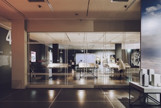 A modern exhibit room with large tables displaying documents and objects, encased in glass. A person is viewing the exhibit. The room features high ceilings and various lighting setups, giving it a contemporary look.