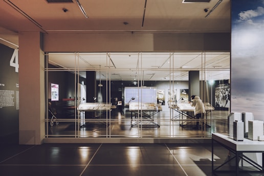 A modern exhibit room with large tables displaying documents and objects, encased in glass. A person is viewing the exhibit. The room features high ceilings and various lighting setups, giving it a contemporary look.