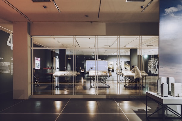A modern exhibit room with large tables displaying documents and objects, encased in glass. A person is viewing the exhibit. The room features high ceilings and various lighting setups, giving it a contemporary look.