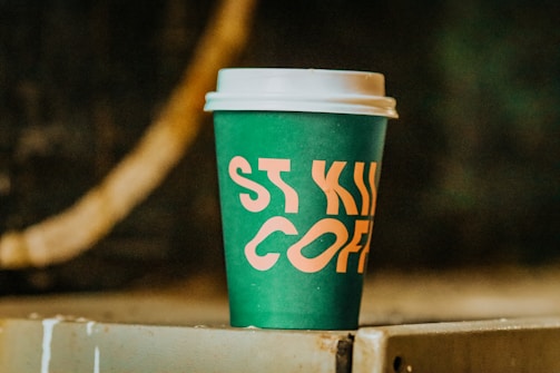 A green disposable coffee cup with a white lid sits on a surface. The cup has bold, orange lettering that partially spells out 'ST KIL COFFEE'. The background is blurred and dark, creating a contrast with the brightly colored cup.