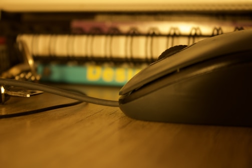 A close-up of a computer mouse on a desk. In the background, there are spiral-bound notebooks with one having a blue cover. The image has a warm, sepia-like tone.