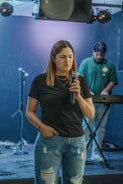 A woman with long hair holds a microphone, looking expressive in a dimly lit room. She wears a black t-shirt and ripped blue jeans. In the background, a man stands at a keyboard, focusing on his equipment. The setup includes microphones on stands and some sound equipment.