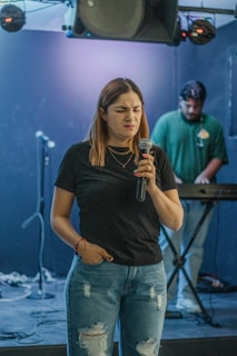 A woman with long hair holds a microphone, looking expressive in a dimly lit room. She wears a black t-shirt and ripped blue jeans. In the background, a man stands at a keyboard, focusing on his equipment. The setup includes microphones on stands and some sound equipment.