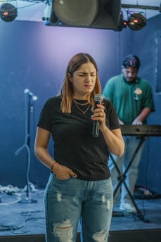 A woman with long hair holds a microphone, looking expressive in a dimly lit room. She wears a black t-shirt and ripped blue jeans. In the background, a man stands at a keyboard, focusing on his equipment. The setup includes microphones on stands and some sound equipment.