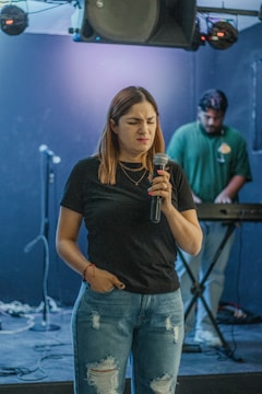 A woman with long hair holds a microphone, looking expressive in a dimly lit room. She wears a black t-shirt and ripped blue jeans. In the background, a man stands at a keyboard, focusing on his equipment. The setup includes microphones on stands and some sound equipment.