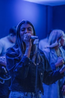 A woman is singing passionately into a microphone while others are present in the background. The lighting is dim with blue hues creating a serene ambiance. She is wearing a denim jacket and appears focused on her performance.
