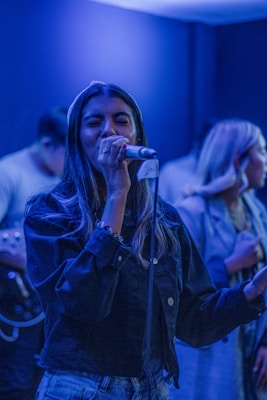 A woman is singing passionately into a microphone while others are present in the background. The lighting is dim with blue hues creating a serene ambiance. She is wearing a denim jacket and appears focused on her performance.