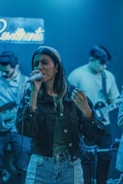 A woman sings passionately into a microphone while two musicians play guitars in the background. The scene is lit with a cool blue light, and a neon sign hangs on the wall.