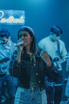 A woman sings passionately into a microphone while two musicians play guitars in the background. The scene is lit with a cool blue light, and a neon sign hangs on the wall.