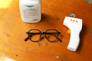 A wooden surface displays three objects: a white bottle with a label, a pair of black glasses, and a white infrared thermometer with orange buttons.