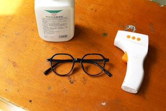 A wooden surface displays three objects: a white bottle with a label, a pair of black glasses, and a white infrared thermometer with orange buttons.