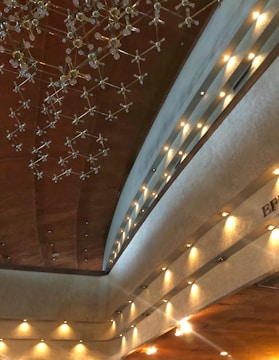 An architectural interior featuring a high ceiling adorned with a modern, artistic chandelier composed of metal and glass. The ceiling and walls are illuminated by numerous small recessed lights, casting a warm glow on the textured surfaces. The overall design is elegant and contemporary.