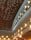 An architectural interior featuring a high ceiling adorned with a modern, artistic chandelier composed of metal and glass. The ceiling and walls are illuminated by numerous small recessed lights, casting a warm glow on the textured surfaces. The overall design is elegant and contemporary.