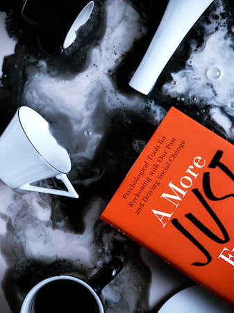 Several coffee cups and a bright orange book are placed on a marbled surface. The cups vary in color, with some being white and others black, and they are filled with black coffee. The book has bold text, suggesting themes of psychology and social change.