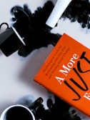 An orange book titled 'A More Just' lies on a surface surrounded by two black coffee cups. Black liquid is spilled around, creating an abstract and dramatic effect.