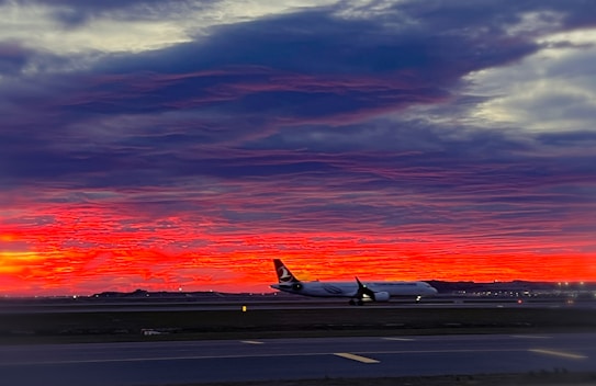 A sleek airplane is on the ground, set against a vivid, dramatic sunset sky with deep red and orange hues. The silhouette of the plane is clearly visible, and the scene captures a moment of tranquility and beauty at an airport. The runway is faintly visible in the foreground as dusk settles over the landscape.