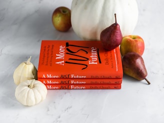 Three stacked books with bright orange covers titled 'A More Just Future' are accompanied by fruits and miniature white pumpkins. The fruits include ripe pears and apples, adding natural elements to the composition.