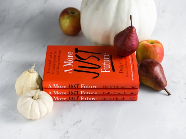 Three stacked books with bright orange covers titled 'A More Just Future' are accompanied by fruits and miniature white pumpkins. The fruits include ripe pears and apples, adding natural elements to the composition.