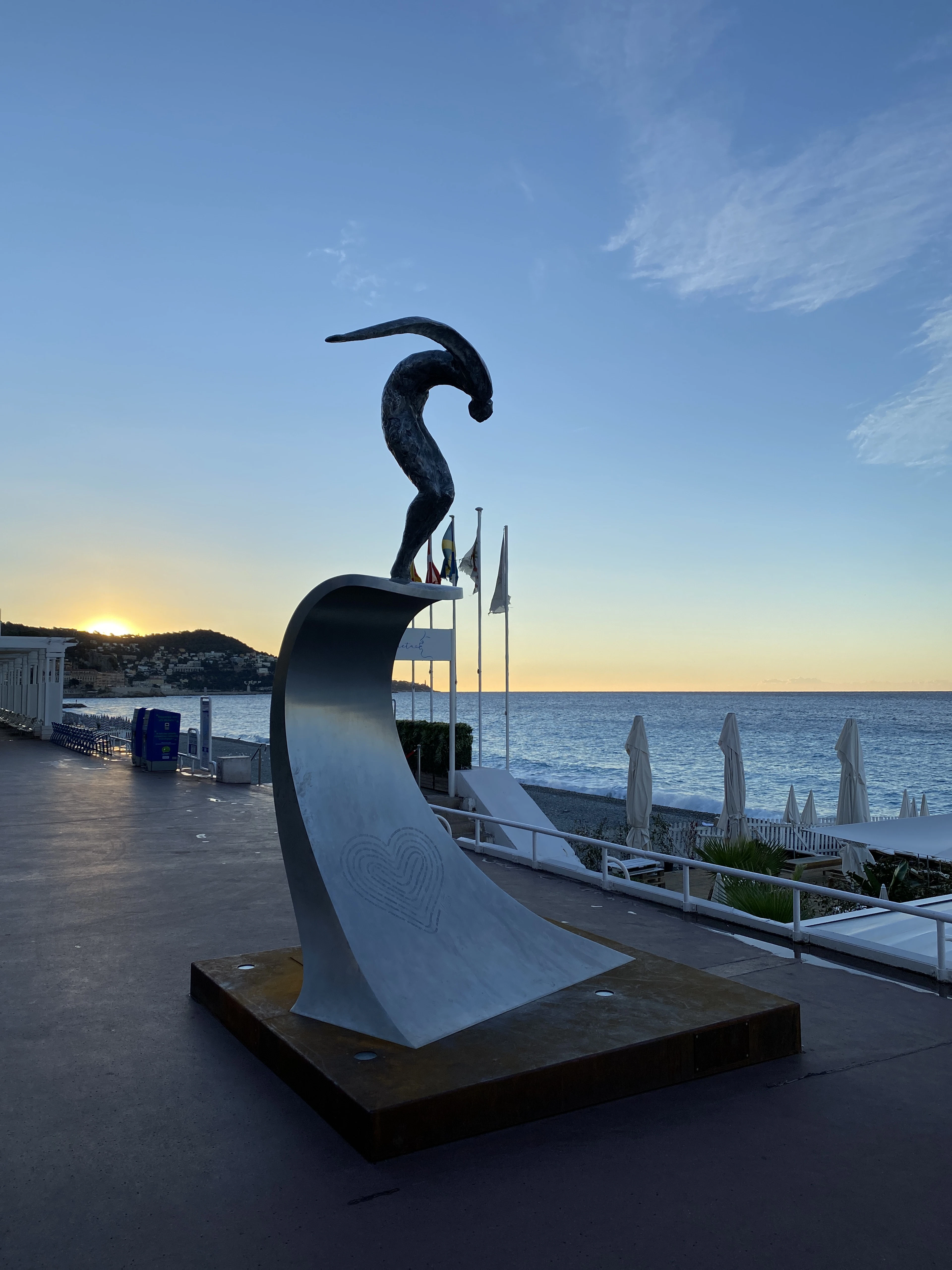 A stainless-steel diving-figure sculpture on the seafront promenade silhouetted at sunset.
