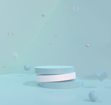 The image depicts a minimalist, abstract composition with several geometric shapes, including a stack of circular platforms, spheres, rings, and a cube, all in shades of light blue and white, floating in a blue-toned environment.