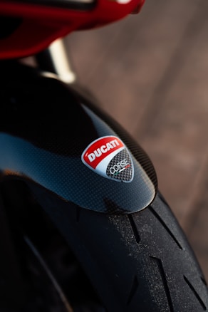 A close-up of a motorcycle tire featuring a shiny black carbon fiber fender with a red and white Ducati logo prominently displayed.