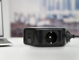 A black power strip is placed on a white surface, featuring a standard socket and several USB charging ports. The background appears to be blurred, likely capturing a workspace or office environment.