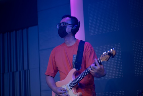A person wearing black-rimmed glasses, a black face mask, and headphones, playing an electric guitar. The individual is dressed in a red T-shirt and appears to be in a music studio or performance space with blue and purple lighting.