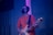 A person wearing black-rimmed glasses, a black face mask, and headphones, playing an electric guitar. The individual is dressed in a red T-shirt and appears to be in a music studio or performance space with blue and purple lighting.