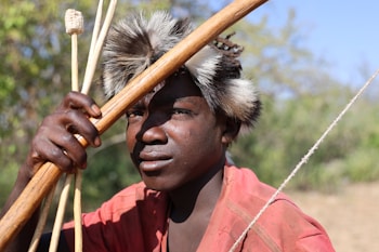 A person wearing a red garment holds a bow and arrows. They have a fur headband. The background is a blurred natural setting with greenery and blue sky.