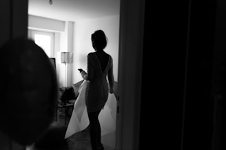A silhouette of a person in a dress holding a phone, standing in a dimly lit room. The room features modern furnishings including a lamp, sofa, and a window letting in natural light.