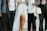 A group of people dressed in formal and semi-formal attire. One figure is wearing a white mesh dress with a long flowing white fabric. Another person is holding a pair of glasses and carrying a patterned bag. The ensemble includes suits, a white shirt, and patterned garments, suggesting a festive or ceremonial occasion.