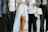 A group of people dressed in formal and semi-formal attire. One figure is wearing a white mesh dress with a long flowing white fabric. Another person is holding a pair of glasses and carrying a patterned bag. The ensemble includes suits, a white shirt, and patterned garments, suggesting a festive or ceremonial occasion.