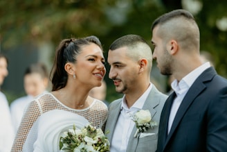 A woman in a white dress with floral details interacts closely with two men wearing suits. They are all smiling and appear to be engaged in a friendly conversation. The background is blurred with greenery, suggesting an outdoor setting.