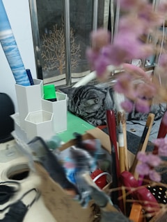 A cozy scene with a cat sleeping amidst various art supplies and stationery items, including brushes, a cardboard box, and containers for organizing tools. A blurred foreground of pinkish flowers adds a soft touch, while a decorative partition in the background shows an image of a tree.