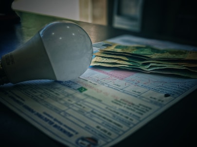 A light bulb is placed on top of a stack of paper currency and paperwork, likely bills or invoices. The scene suggests a focus on electricity costs, with soft lighting creating shadows.