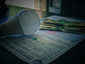 A light bulb is placed on top of a stack of paper currency and paperwork, likely bills or invoices. The scene suggests a focus on electricity costs, with soft lighting creating shadows.