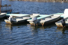 Several small motorboats are moored at a dock on a body of water. The boats are lined up neatly, and each has an outboard motor attached. The water appears calm, and the dock has a weathered appearance with a light blue-green wooden surface.