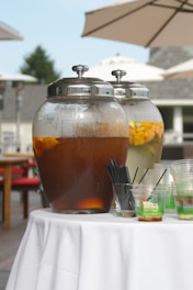 Large glass beverage dispensers filled with drinks stand on a white tablecloth under outdoor umbrellas. One dispenser contains a dark liquid, possibly iced tea, with orange slices, while the other holds a lighter drink with floating fruit pieces, suggesting infused water.