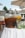 Large glass beverage dispensers filled with drinks stand on a white tablecloth under outdoor umbrellas. One dispenser contains a dark liquid, possibly iced tea, with orange slices, while the other holds a lighter drink with floating fruit pieces, suggesting infused water.