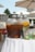 Large glass beverage dispensers filled with drinks stand on a white tablecloth under outdoor umbrellas. One dispenser contains a dark liquid, possibly iced tea, with orange slices, while the other holds a lighter drink with floating fruit pieces, suggesting infused water.