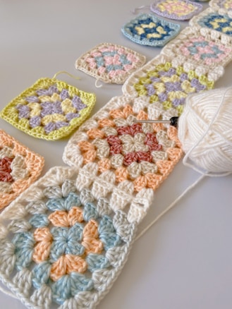 A collection of crocheted granny squares in various pastel colors are arranged on a flat surface. A crochet hook is inserted into one of the squares, and a ball of white yarn is nearby. The granny squares are linked together in a chain and include colors such as light blue, peach, lavender, pink, and yellow.