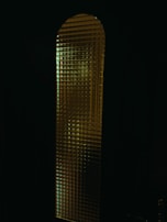 A tall, narrow glass window with a textured surface that distorts the light passing through. The glass pattern seems to be grid-like, creating a mosaic effect. The predominantly dark surroundings make the illuminated window stand out more, giving a mysterious vibe.