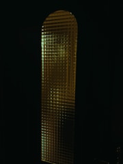 A tall, narrow glass window with a textured surface that distorts the light passing through. The glass pattern seems to be grid-like, creating a mosaic effect. The predominantly dark surroundings make the illuminated window stand out more, giving a mysterious vibe.