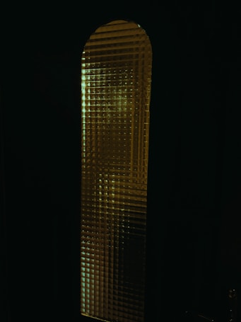 A tall, narrow glass window with a textured surface that distorts the light passing through. The glass pattern seems to be grid-like, creating a mosaic effect. The predominantly dark surroundings make the illuminated window stand out more, giving a mysterious vibe.