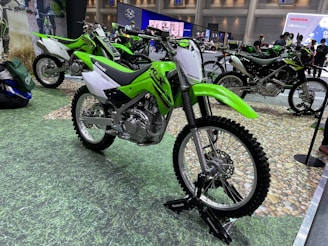 A display of several Kawasaki dirt bikes with vibrant green and white colors, showcased in a well-lit exhibition space. The bikes are positioned on a textured floor resembling grass and gravel, with banners and advertisements from other brands visible in the background.