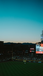 A baseball stadium during a game is captured with a scoreboard in the background displaying player information. The sky features a gradient transitioning from blue at the top to orange near the horizon as the sun sets. The field is partially visible with a player discernible near the outfield, and the stands are filled with spectators.